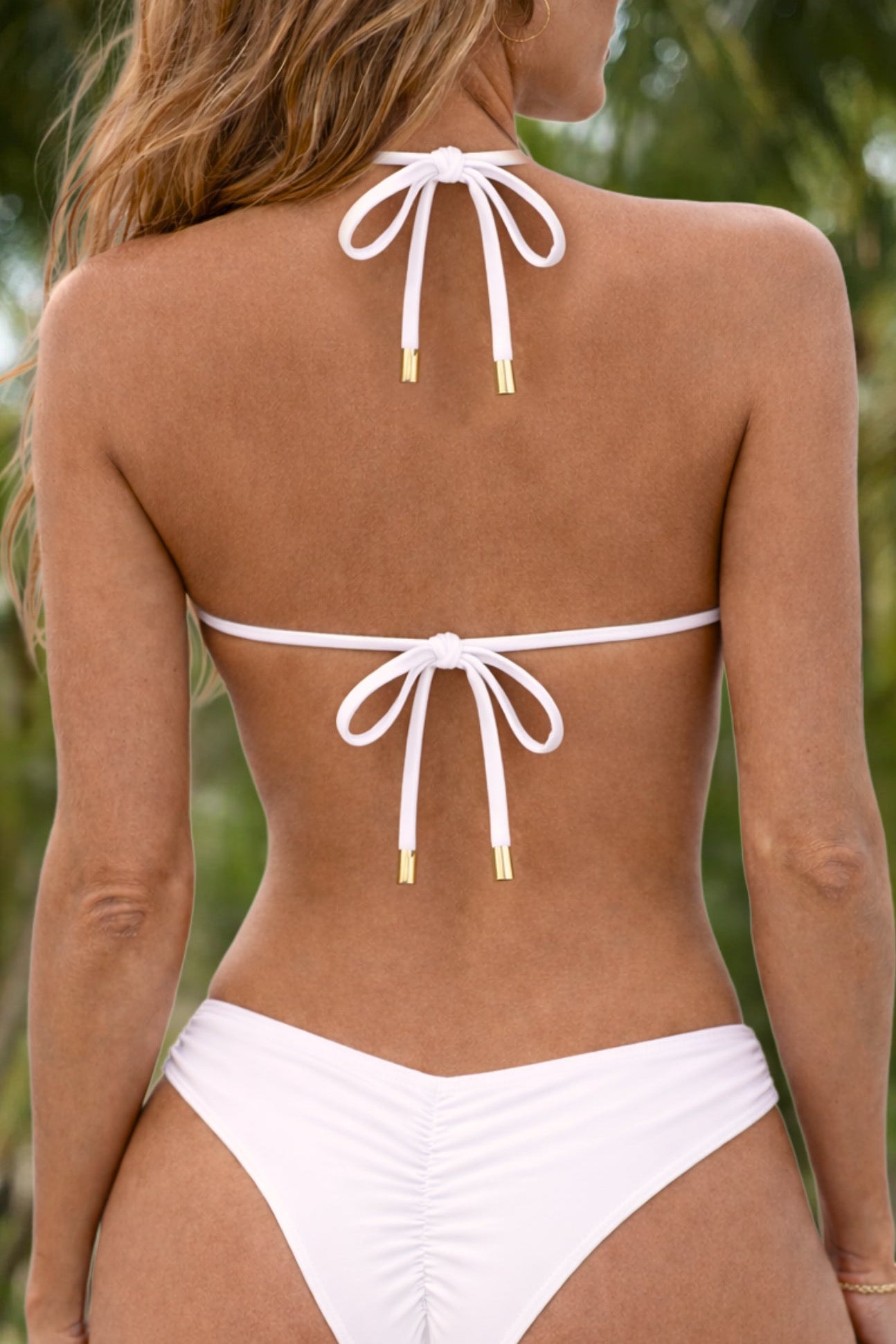 White string bikini bottom and matching top shown from the back on a model, featuring gold-toned tip details on the tie strings, with a blurred green outdoor background.